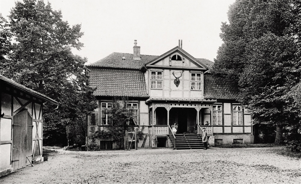 Zu sehen ist ein Fachwerkhaus mit Vortreppe und Veranda. Auf der Veranda sitzen zwei Frauen, vor der Treppe steht der Förster.